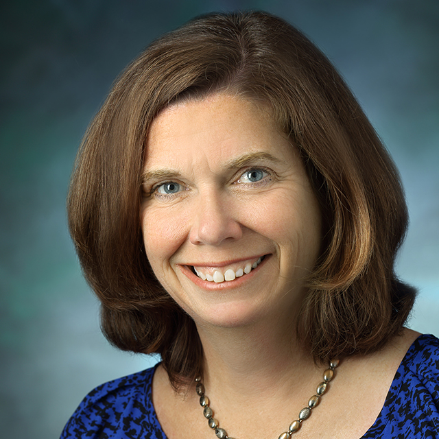 Sue De Pasquale smiles and stands against a blue-grey background. 